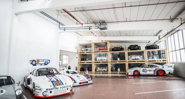 Spacious garage showcasing a collection of classic Martini Porsche race cars, with sleek designs and iconic liveries, strategically positioned for display and set against a backdrop of neatly arranged automotive memorabilia on wooden shelves